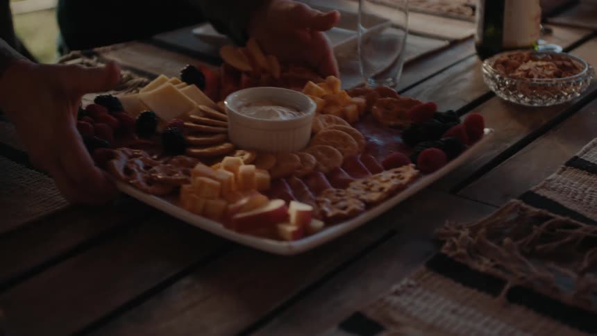 An adult puts a tray of fruit and cheese appetizers on top of an outdoor wooden table