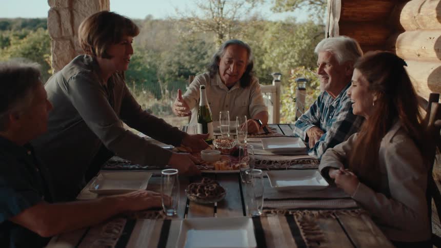 Friends smile when their host serves the meal during a party on the porch of a rural home