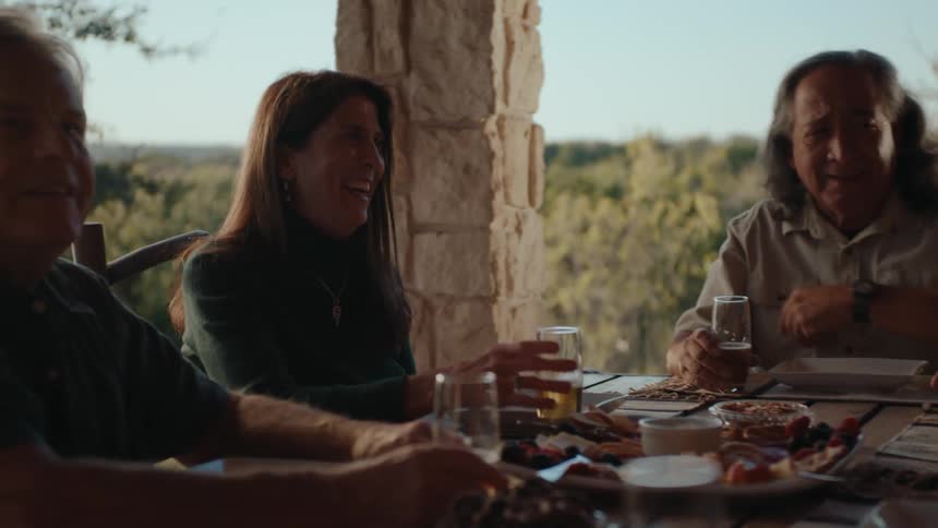 People seated at a table outdoors with plates glasses food wine and vegetation in the background