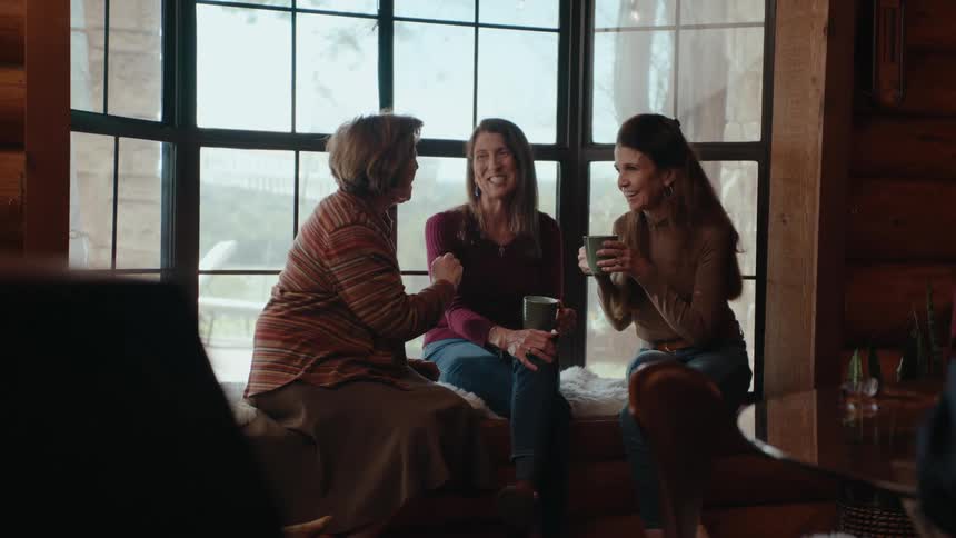 Three women are seated near a large window laughing and talking while holding cups