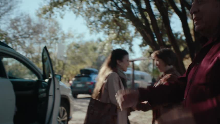 A grayhaired man comes out from behind a car with the trunk open and he is carrying the luggage then greets a man and behind them are two women who are happy to see each other and holding each others hands