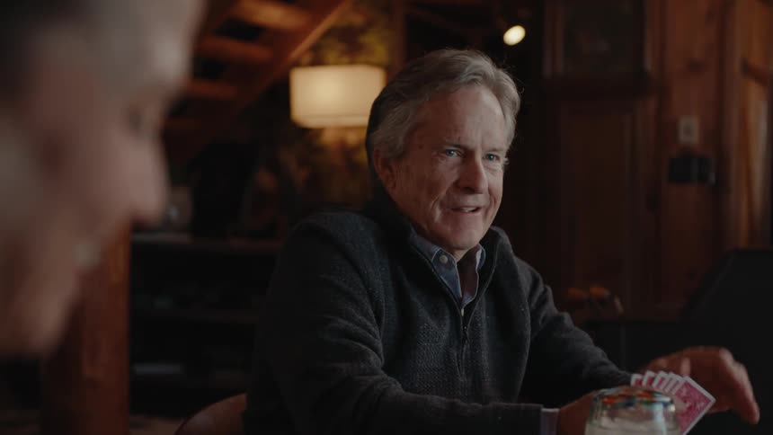 A man plays a game of playing cards smiling in a cozy cabin setting
