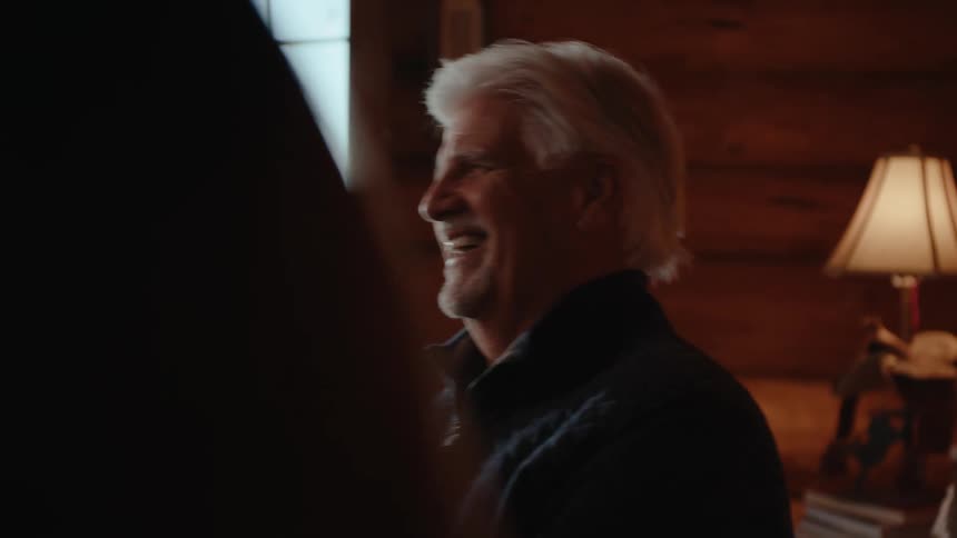 A close up of an older man with a happy expression seated in a log cabin and holding playing cards