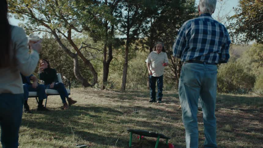 Two old men play a pitching game while a woman brings a drink outside from the house