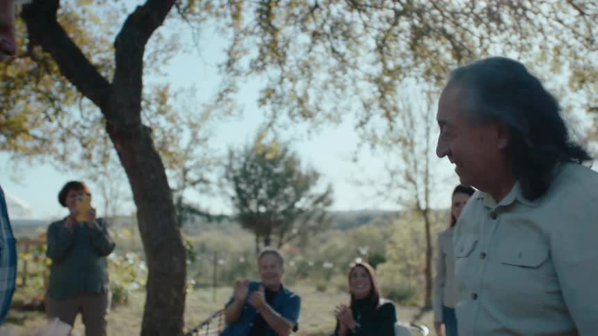 Two elderly men greeting each other and the family next to them applauding while standing in a backyard with trees and mountains
