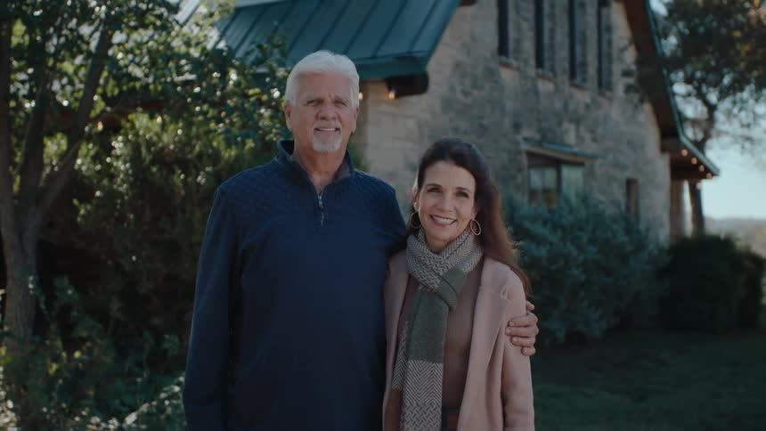 A man and a woman stand in front of a stone house smiling and looking at the camera
