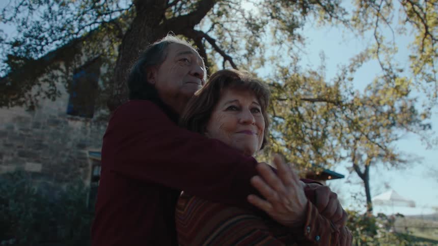 Two older people hug while standing in the yard of a rural house