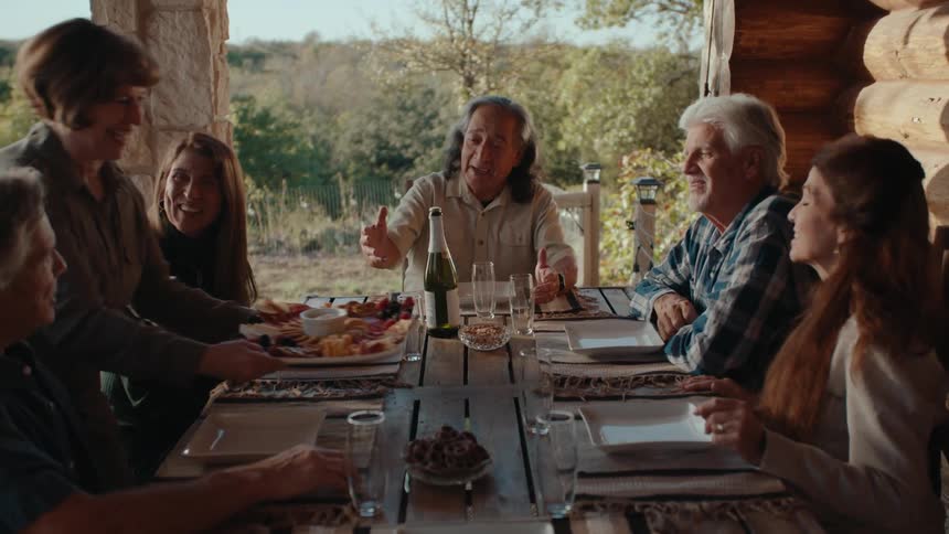 People seated at a table outdoors with plates, glasses, food, wine, and vegetation in the background