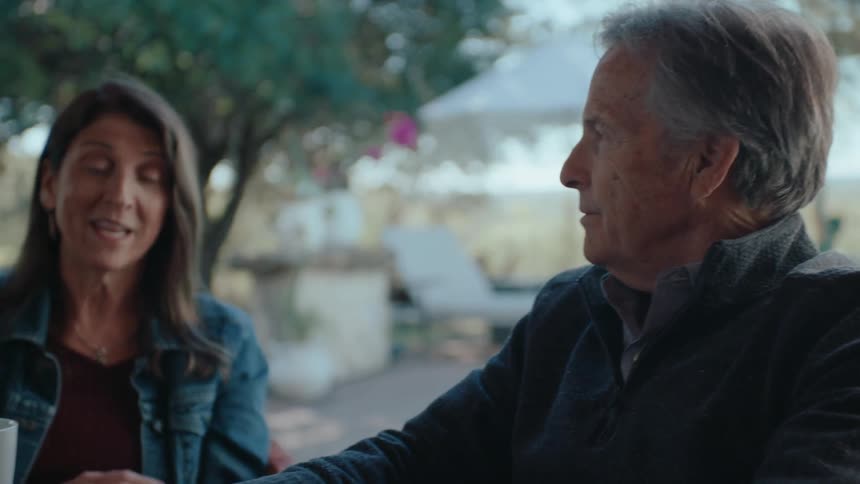 A woman and a man sit at a table outdoors holding mugs while engaged in conversation