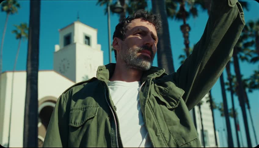 A man with a gray beard and a green jacket looking forward and raising his arm up and behind him some trees and a white building