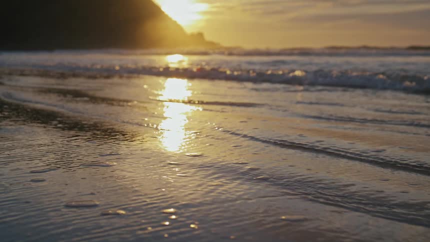 Sea waves breaking at a beach next to a promontory with the sun on the background during dusk or dawn