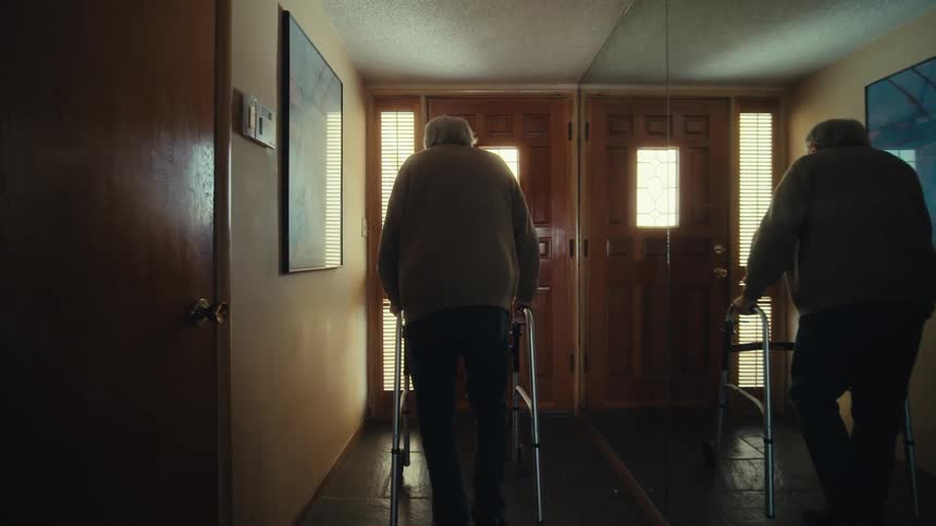 An elderly man is standing with his back turned using a walker to walk down a hallway to a wooden door and to the right of him is a wall of mirrors that reflects him