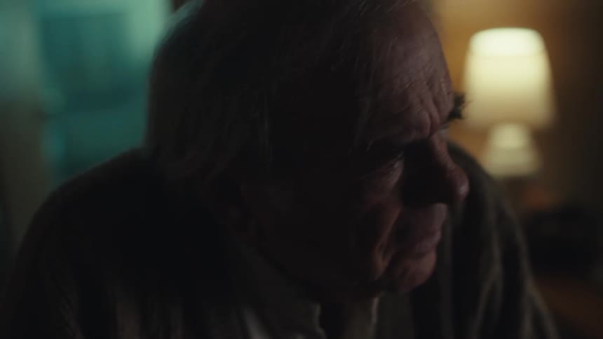 An elderly man wearing a sweater walks into a dimly lit room looking confused