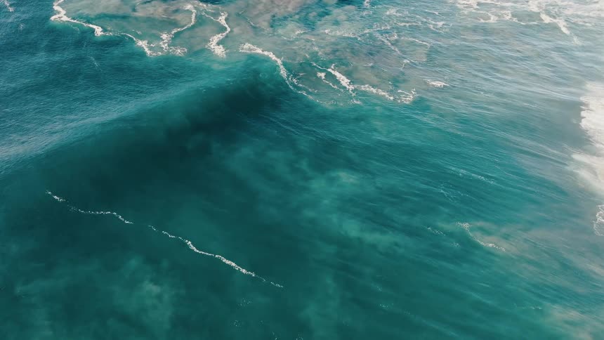 An aerial view of ocean waves crashing onto a shoreline with varying shades of blue and white foam