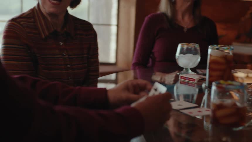 A woman with a smile on her face is passing a playing card to a person in front of her and everyone is sitting around a table full of drinks and cards on the table