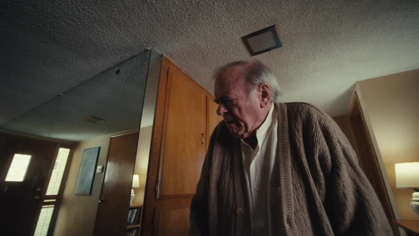 An elderly man wearing a cardigan wanders confusedly around his house looking sad