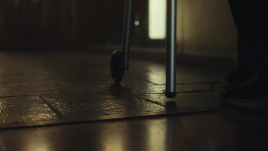 The legs of an elderly man using a walker are visible in the foreground of a dimly lit hallway