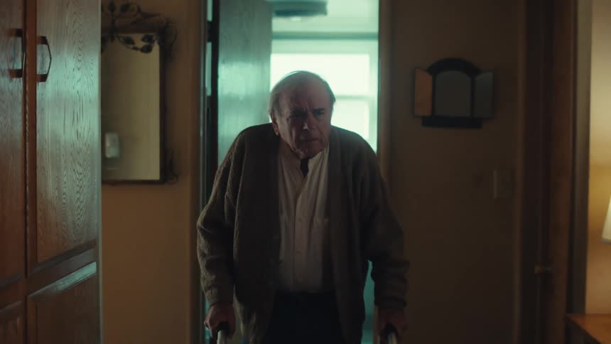 Elderly white man indoors walking with assistance of a walking aid coming out of a bathroom with while frowning and suffering