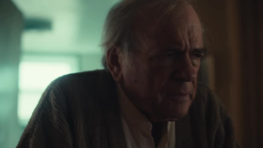 An elderly man wearing a cardigan wanders confusedly around his house looking sad