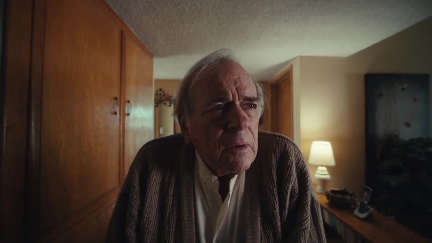 An elderly man wearing a sweater walks into a room looking confused