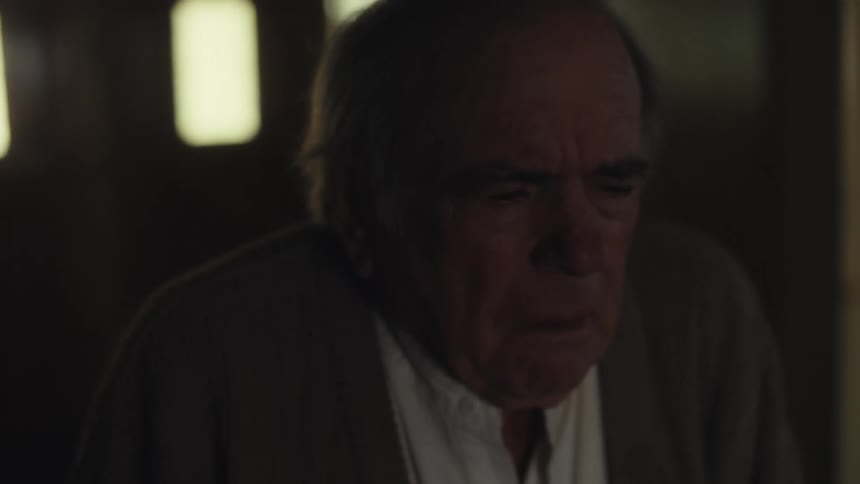 An elderly man stands in a dimly lit room with a tired expression