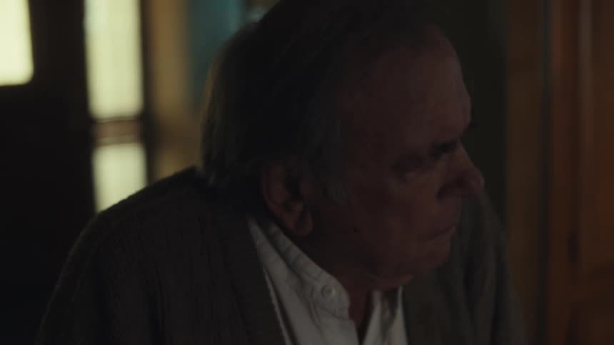 An elderly man wearing a sweater walks into a dimly lit room looking confused