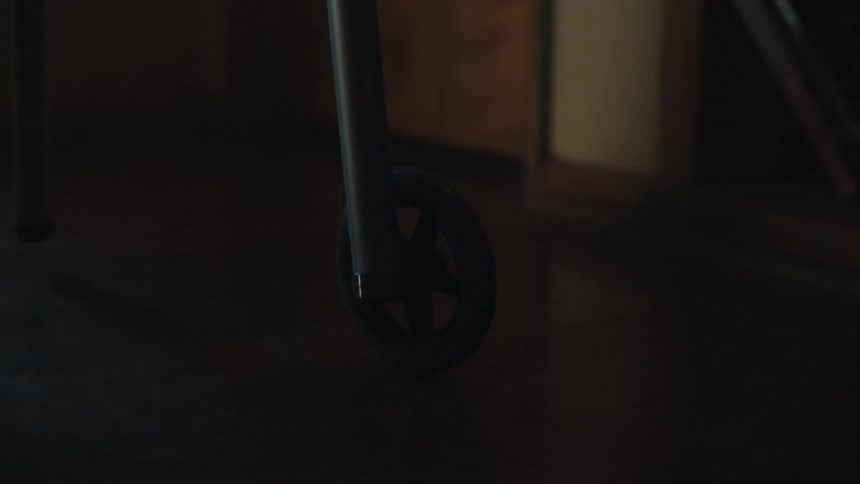 The small black wheel of a walker that is attached to a metal bar and rotating on a smooth textured floor