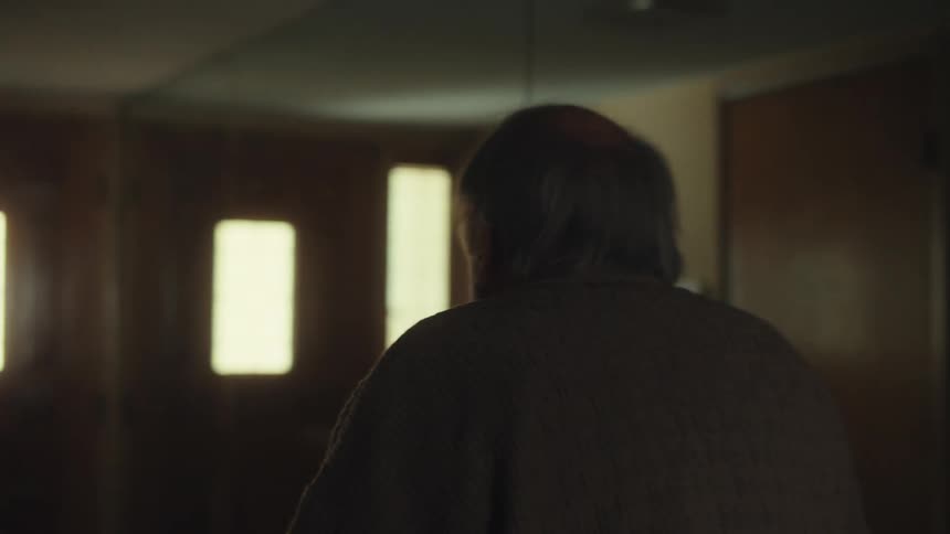 An elderly man wearing a cardigan wanders confusedly around his house looking sad