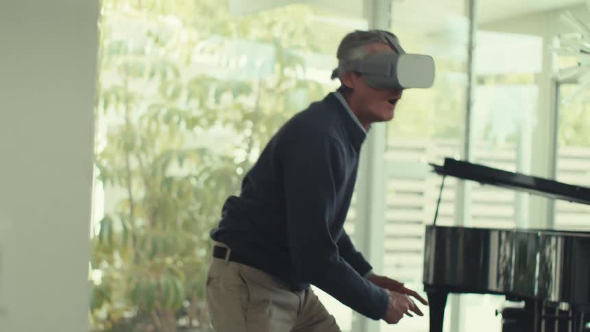 A person is using a virtual reality headset and controllers in a modern sunlit room with a grand piano and contemporary decor
