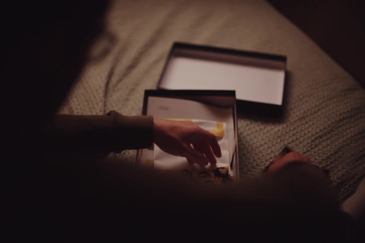 Woman sitting down on a bed looking at a collection of photographs from a black box in front of her indoors