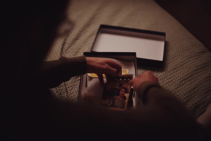 A person is sitting on a bed holding and looking at photographs from an open box