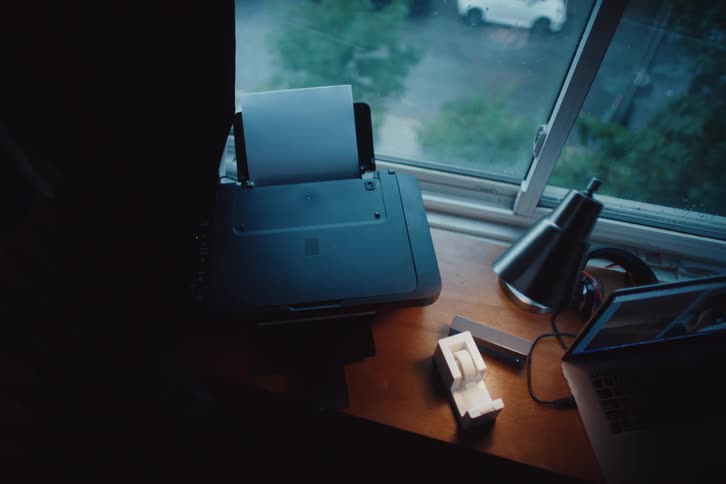 A small printer is printing a document on a desk next to a laptop and a window with rain falling outside