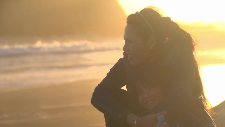 Woman sitting and hugging her child while smiling on a beach at sunset