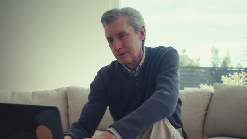 Man smiles and types on a laptop while sitting on a couch