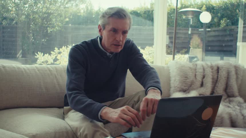 Older man leaning back while viewing laptop in his living room sits up to focus on the screen