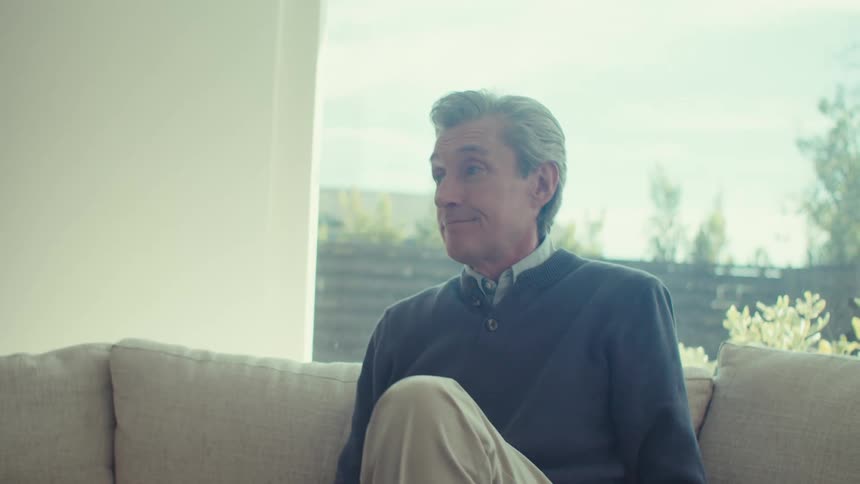 A grayhaired man in a blue coat is sitting on a sofa and he closes a laptop that is in front of him and then stands up