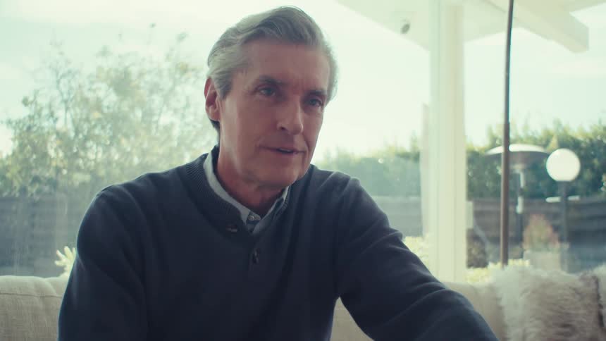Older man sitting on a couch near the window looks up from a laptop and smiles