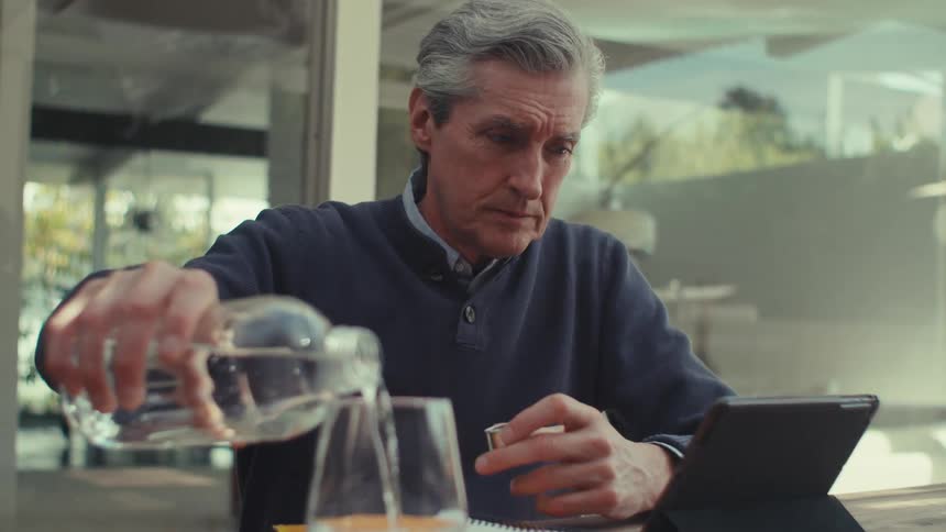 White man sitting at a table pouring himself water from a bottle and looking at the screen of a tablet with a concerned and nervous facial expression during daytime