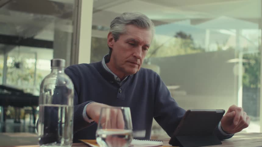 A man is sitting and tapping on a tablet with his finger and then he picks up a pen and starts writing on a piece of paper
