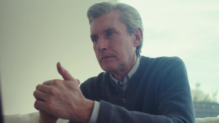 An individual is seated indoors wearing a dark sweater over a collared shirt with hands clasped together
