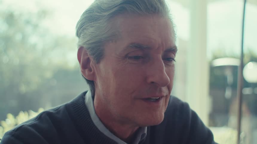 A middle aged man with graying hair sitting indoors near a window engaged in conversation