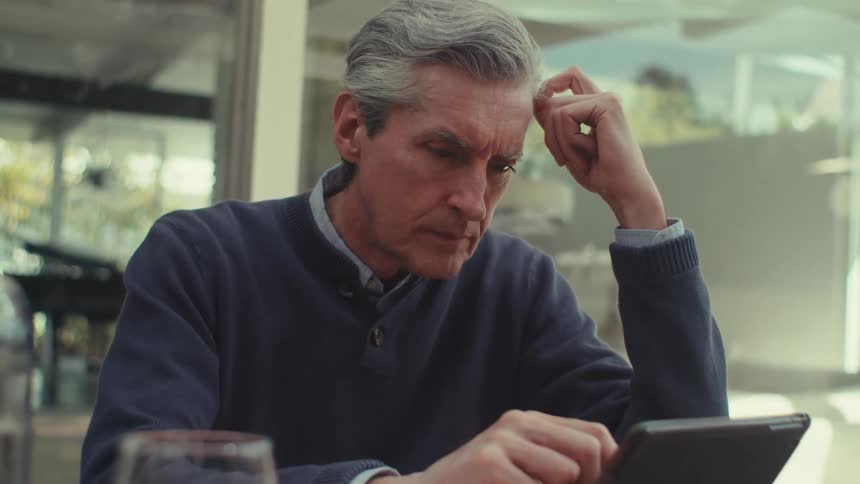 White man sitting down at a table reading from a laptop computer and writing on a notebook next to him during daytime