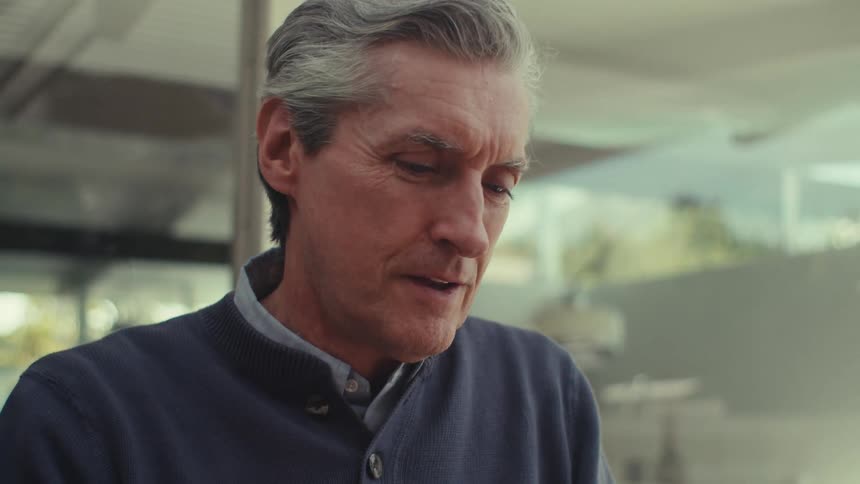 A gray haired man is sitting while tapping a tablet with his finger and he is wearing a subtle smile