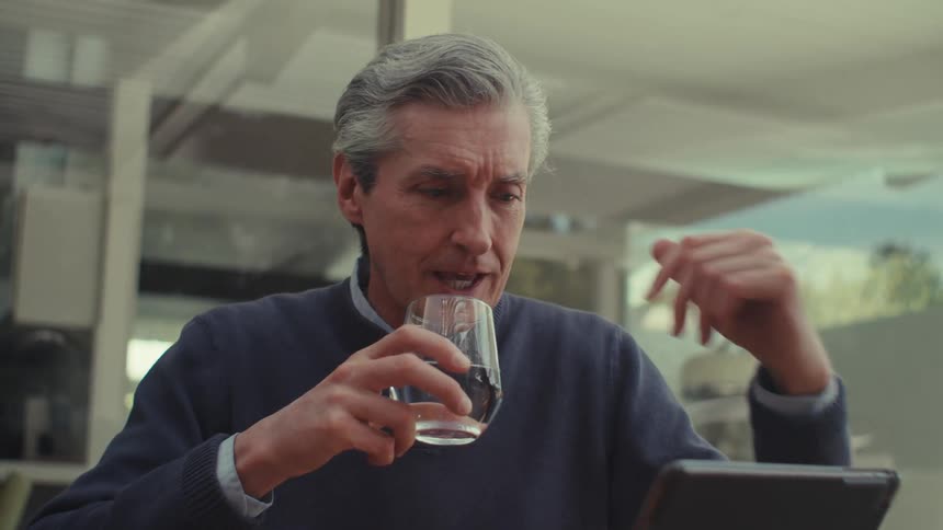 An older individual with gray hair is seated at a table holding a glass of wine and using a tablet in a modern house