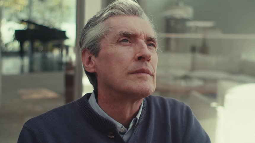 A thoughtful older man sits outdoors looking upward and off into the distance