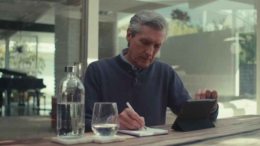 A person is sitting at a wooden table writing in a notebook with a pen while using a tablet with a glass of water and a bottle of water on the table