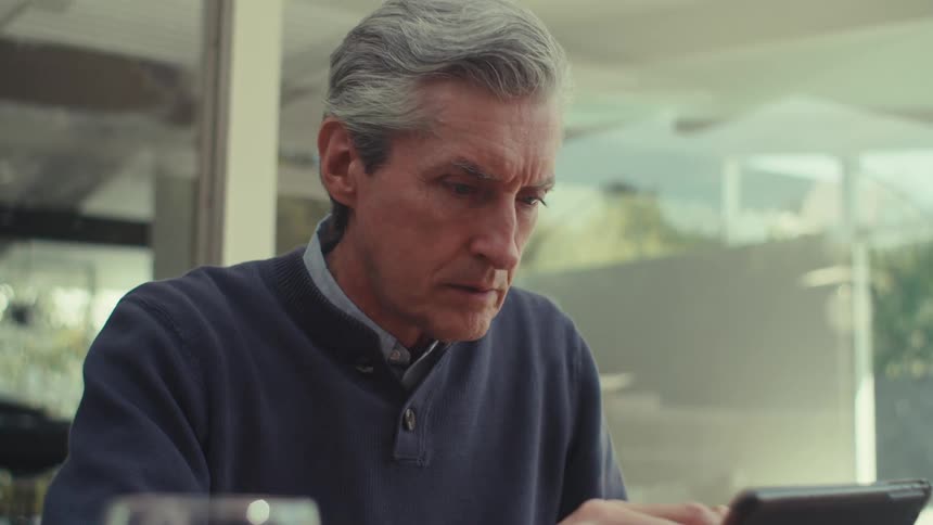 A grayhaired man is fiddling with a tablet in front of him and he is worried