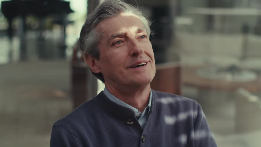A face of a grayhaired man who is sitting in a chair and he is giving a subtle smile and then changes his expression to worried and puts his hands on his face