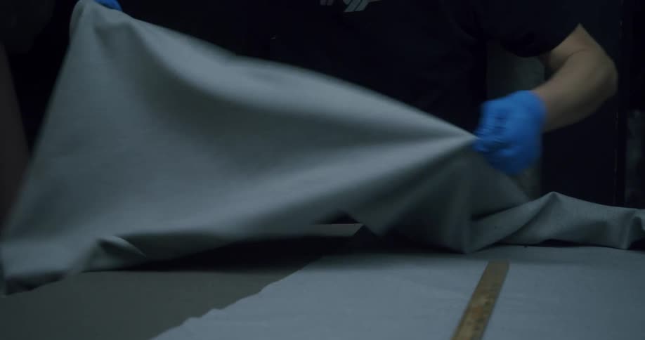 The hands of a person wearing blue gloves handling a gray fabric that is on a table