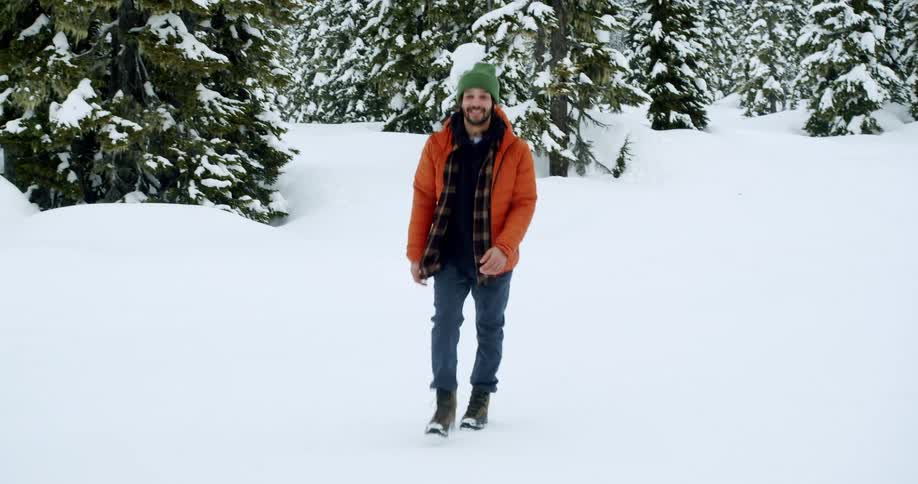Man in an orange jacket walking through a snowy area with conifers in the background